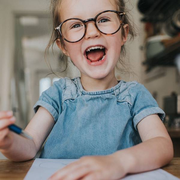 Ein kleines Mädchen mit großer runder Brille, Jeanskleid und einem Stift in der Hand, lacht mit offenem Mund.