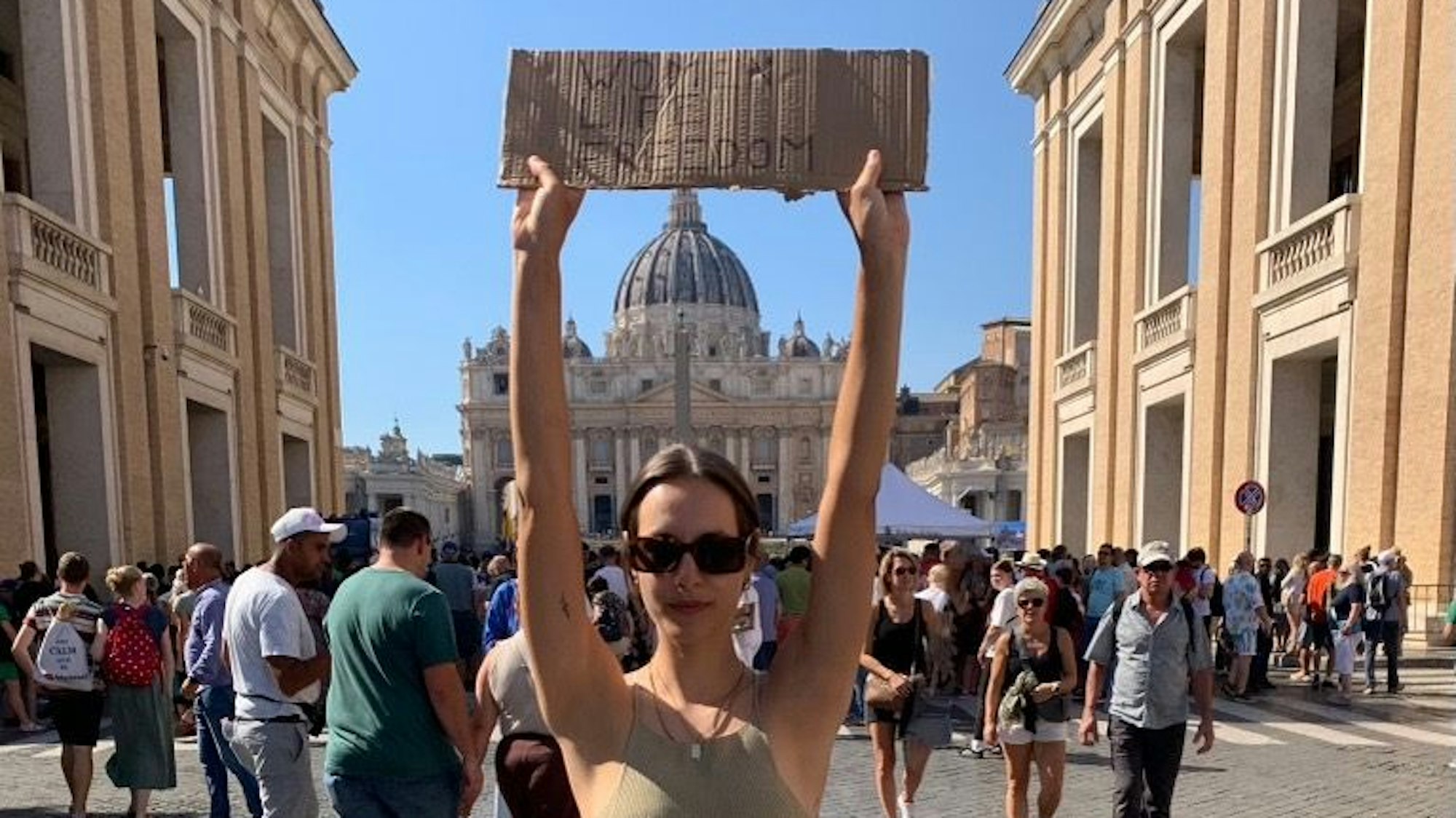 Zu sehen ist Ida Haas. Sie hält ein Schild mit dem Slogan „Woman.Life.Freedom“ in den Händen vor dem Petersdom in Rom.
