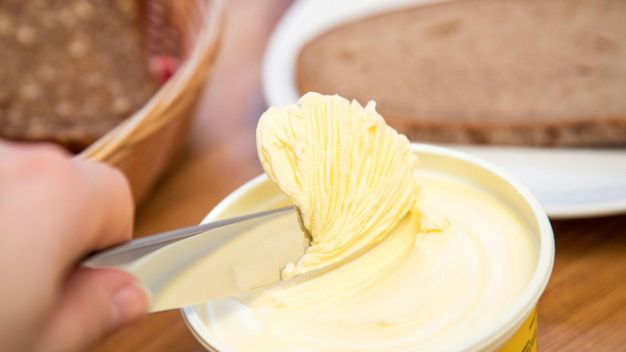 Eine Hand führt ein Messer in einen Topf Margarine, im Hintergrund liegt eine Brotscheibe auf einem Teller.