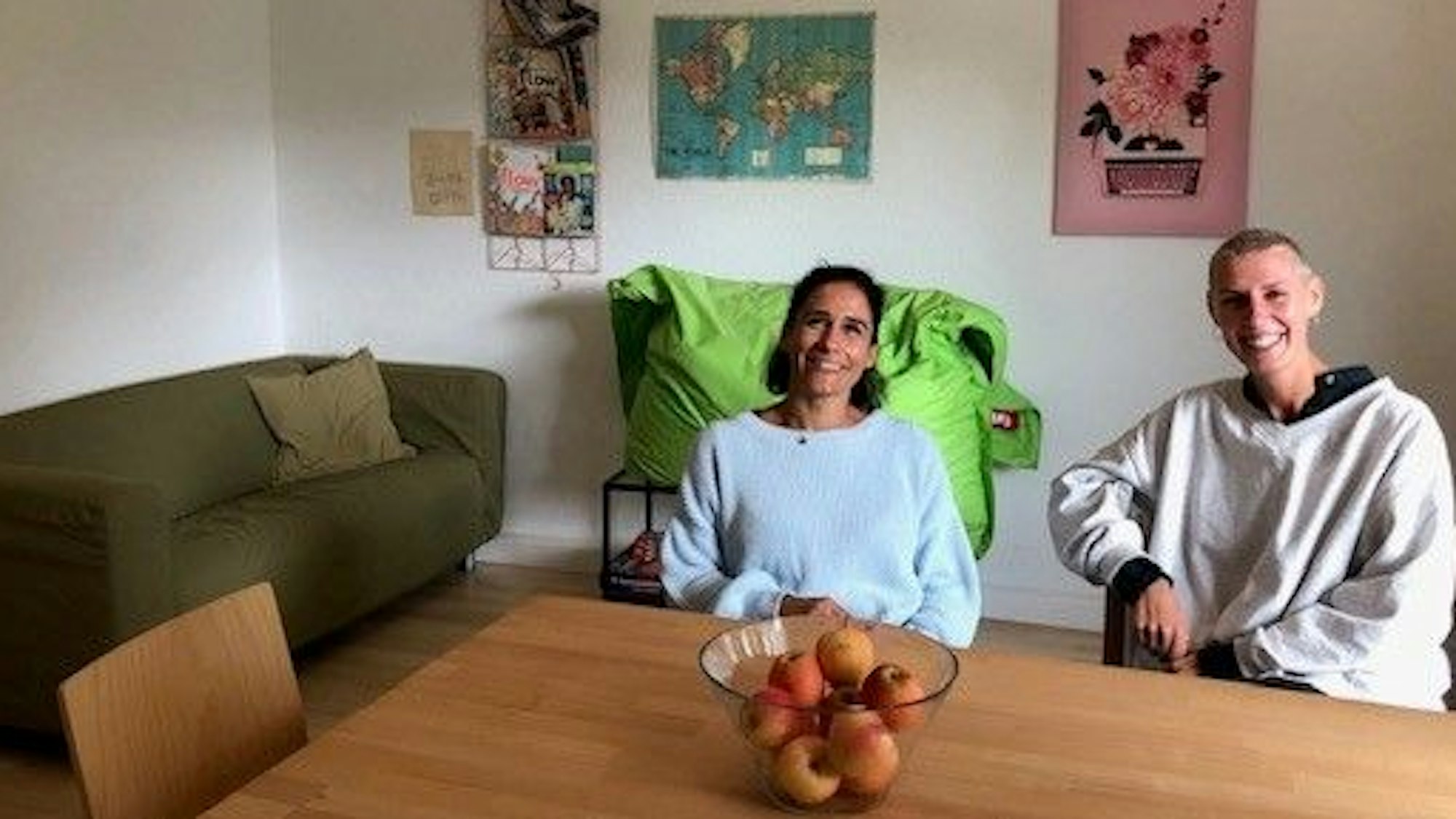 Zwei Mitarbeiterinnen der Notschlafstelle sitzen in dem Wohnzimmer an einem großen Holztisch, im Hintergrund sind ein grünes Sofa und ein froschgrüner Sitzsack zu sehen.