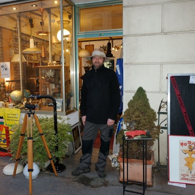 Ein Mann mit Hut und Bart steht vor der Tür eines Ladenlokals, in dem allerlei Lampen und antike Objekte zu sehen sind.