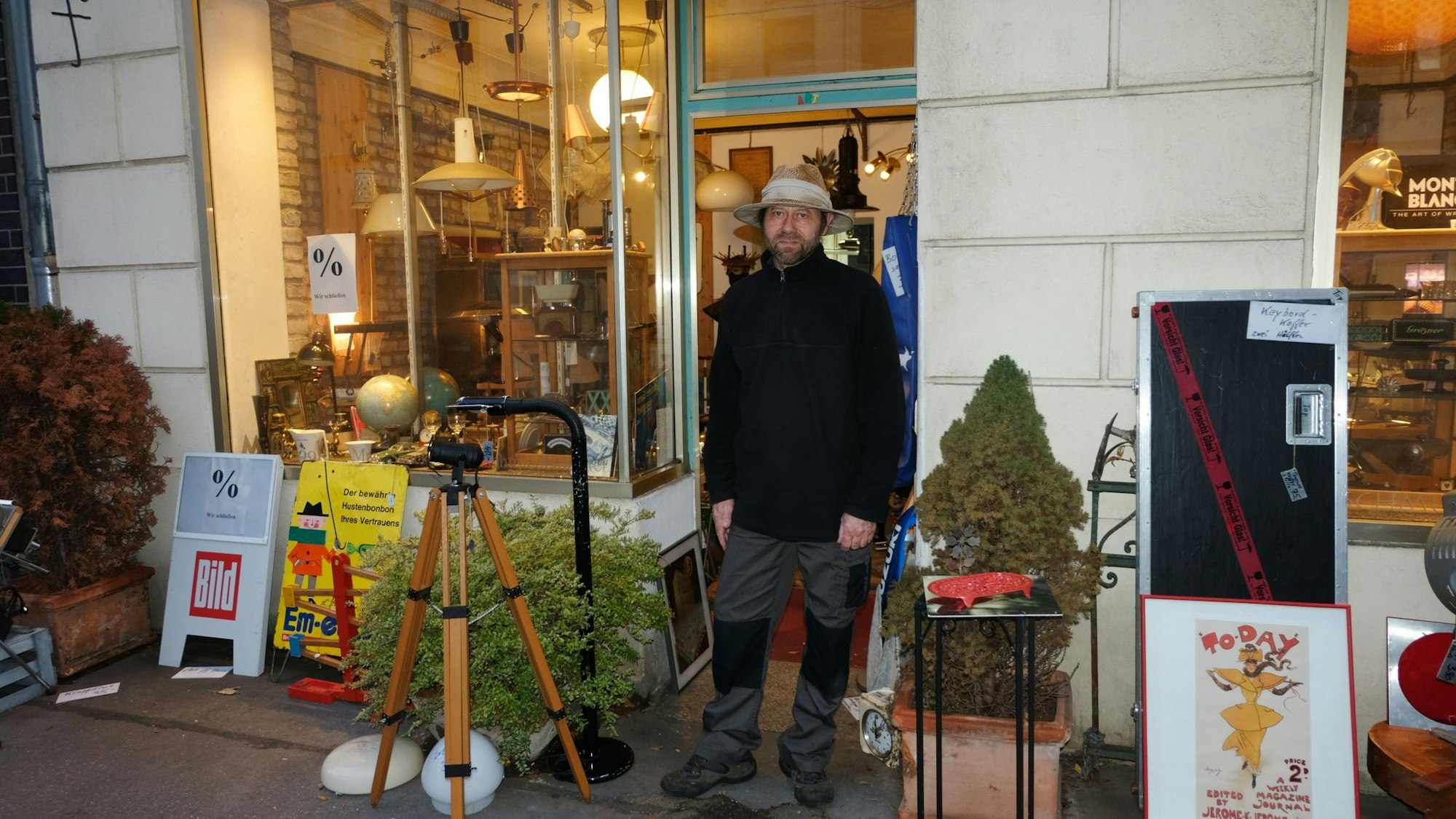 Ein Mann mit Hut und Bart steht vor der Tür eines Ladenlokals, in dem allerlei Lampen und antike Objekte zu sehen sind.