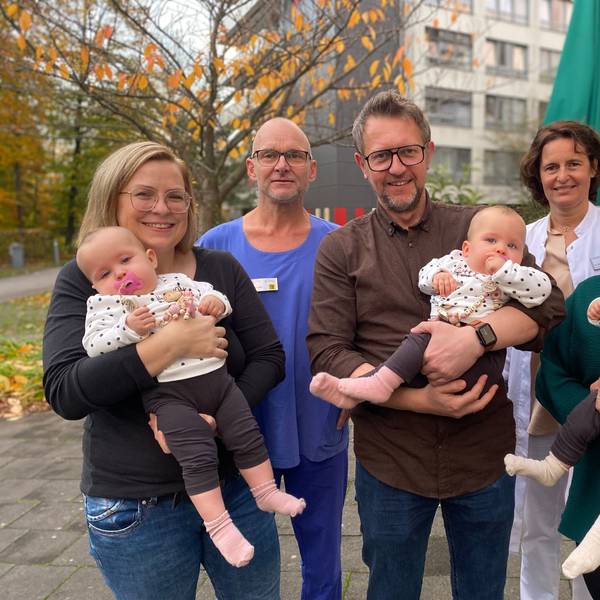 Im Vordergrund stehen zwei Frauen und ein Mann mit jeweils einem Säugling auf dem Arm. Dahinter ein Arzt und eine Ärztin. Im Hintergrund das Klinikum und ein Laubbaum.