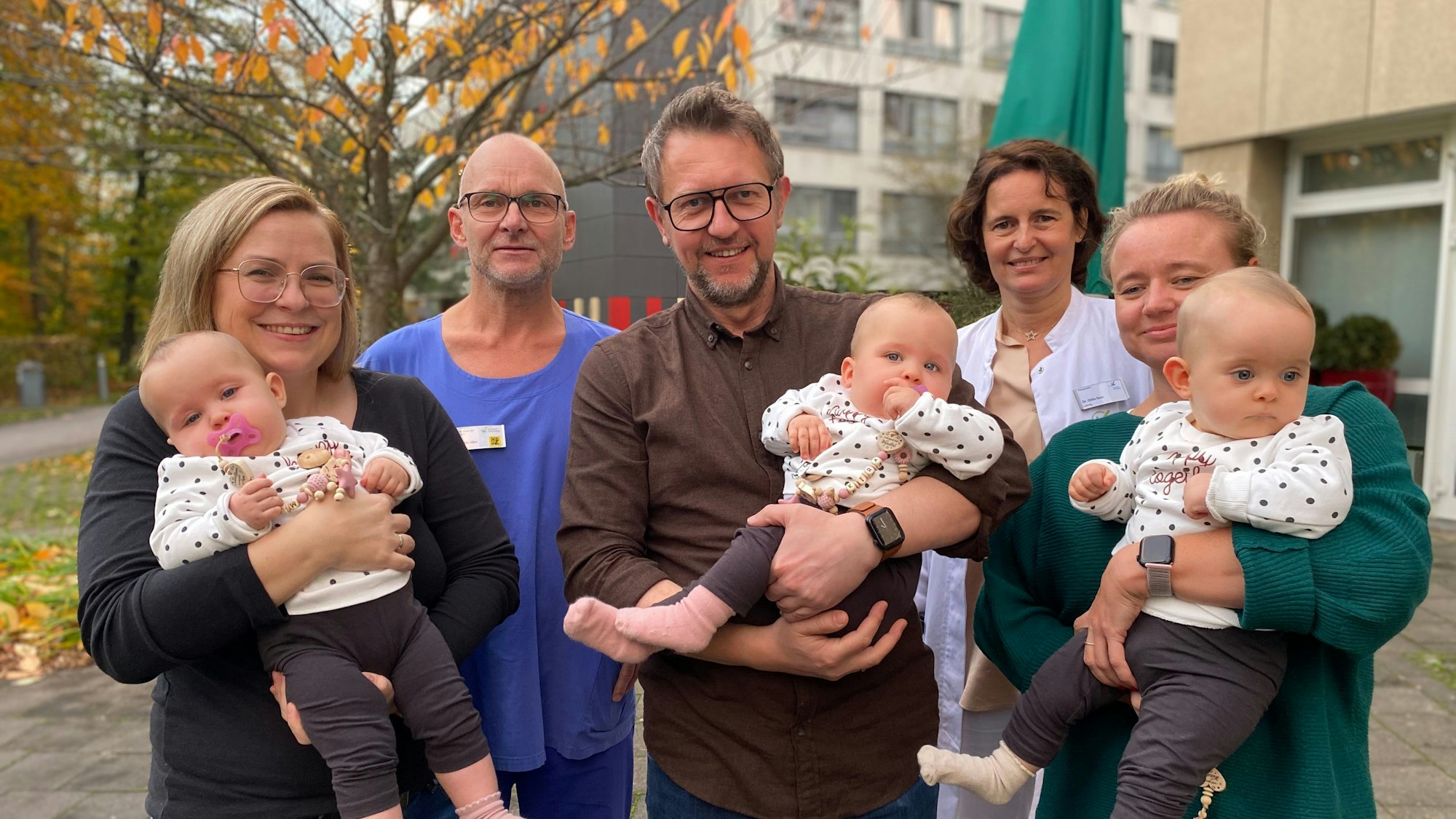 Im Vordergrund stehen zwei Frauen und ein Mann mit jeweils einem Säugling auf dem Arm. Dahinter ein Arzt und eine Ärztin. Im Hintergrund das Klinikum und ein Laubbaum.