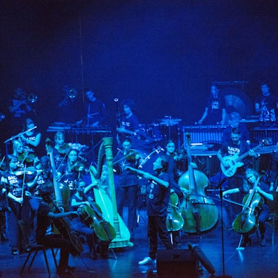 Die Bühne ist dunkel und bläulich erleuchtet. Darauf ist das Orchester, in der Mitte ein Dirigent.