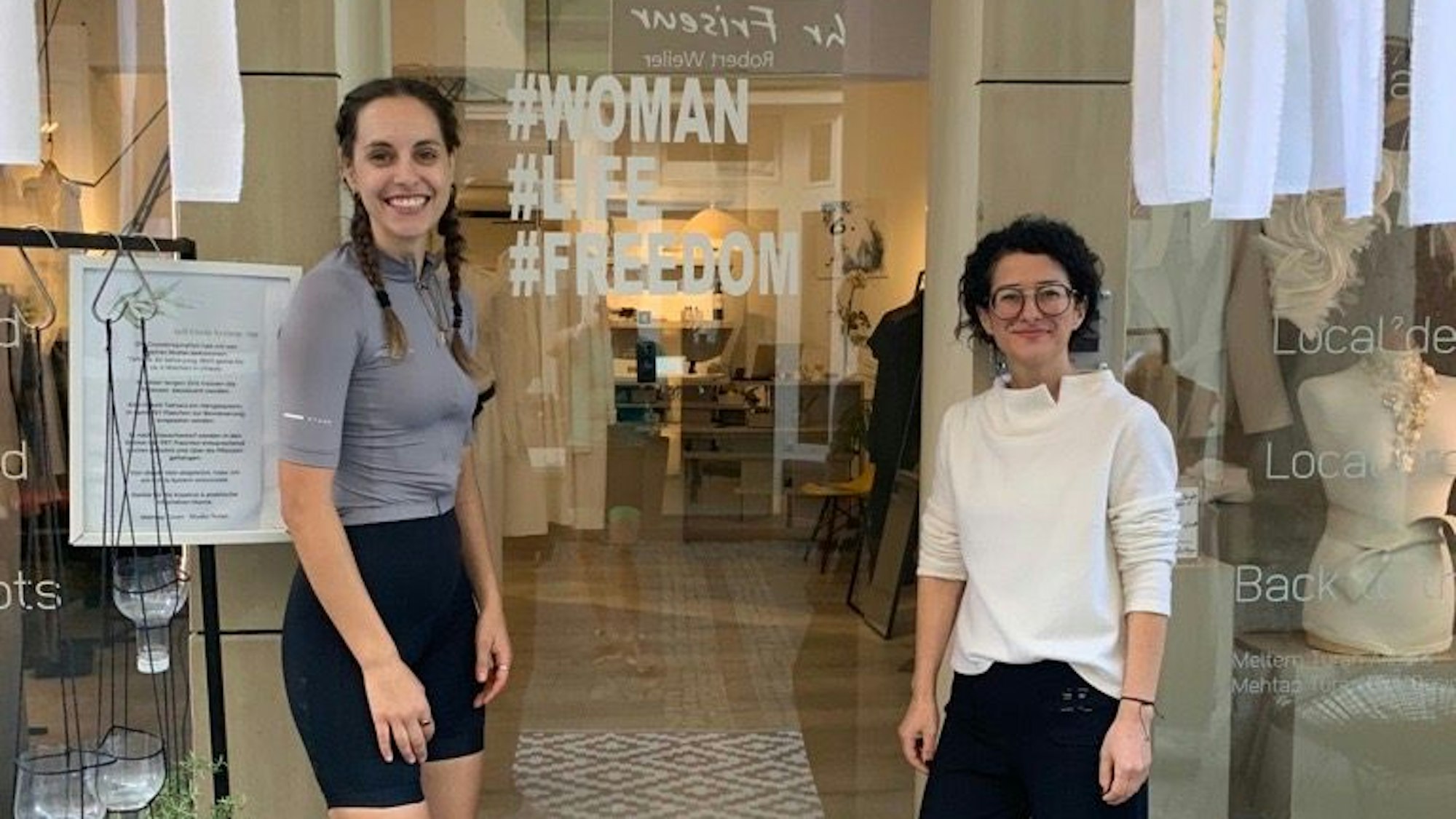 Zu sehen ist die Kölnerin Ida Haas vor einem Modegeschäft. Auf der Fensterfront ist der Revolutionsslogan „Woman.Life.Freedom“ zu sehen.