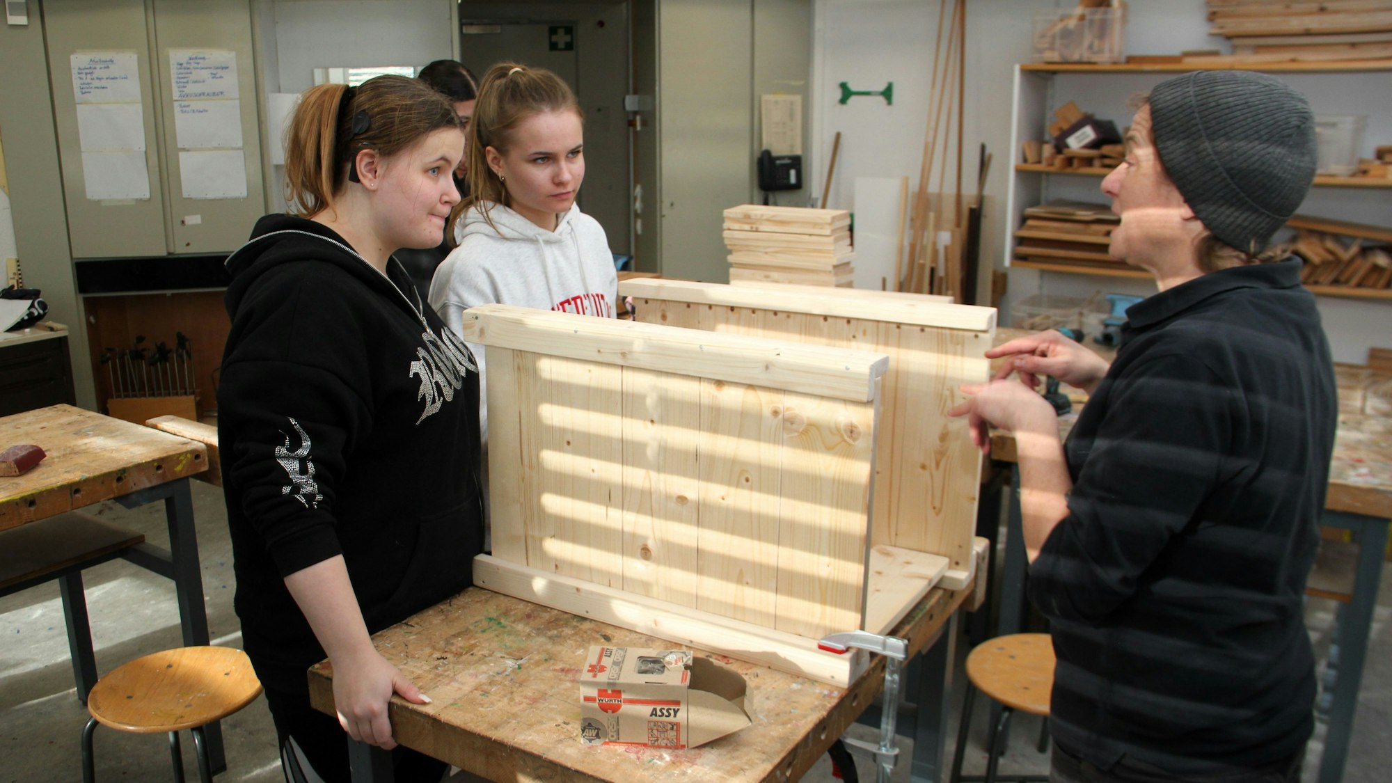 Zwei Schülerinnen und eine Frau mit Mütze werkeln zusammen an einem Möbelstück.