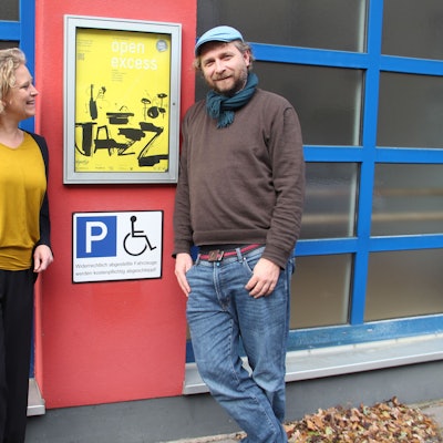 Eine Frau und ein Mann stehen neben einem Veranstaltungsschild und einem Piktogramm Behindertenparkplatz.