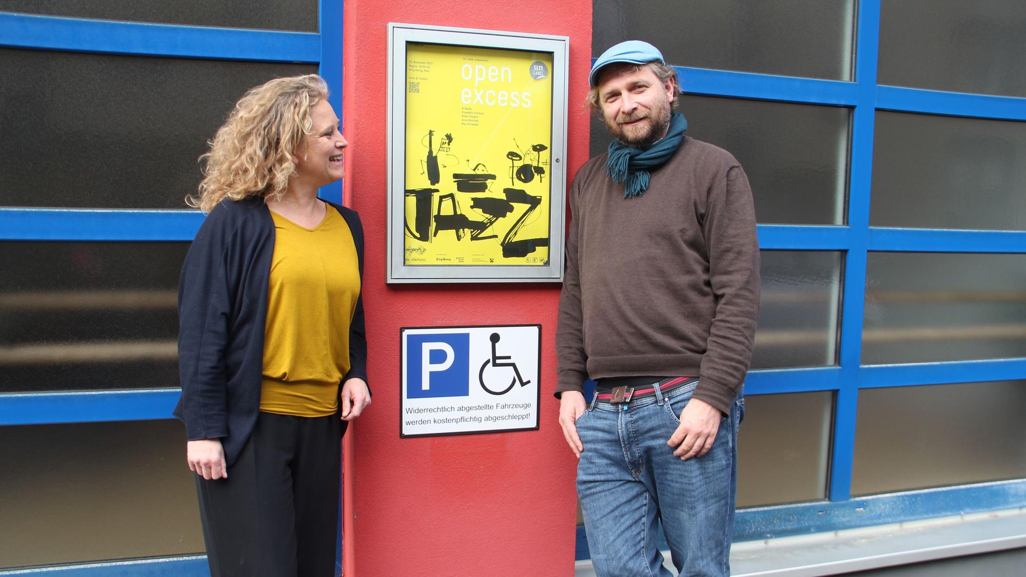 Eine Frau und ein Mann stehen neben einem Veranstaltungsschild und einem Piktogramm Behindertenparkplatz.