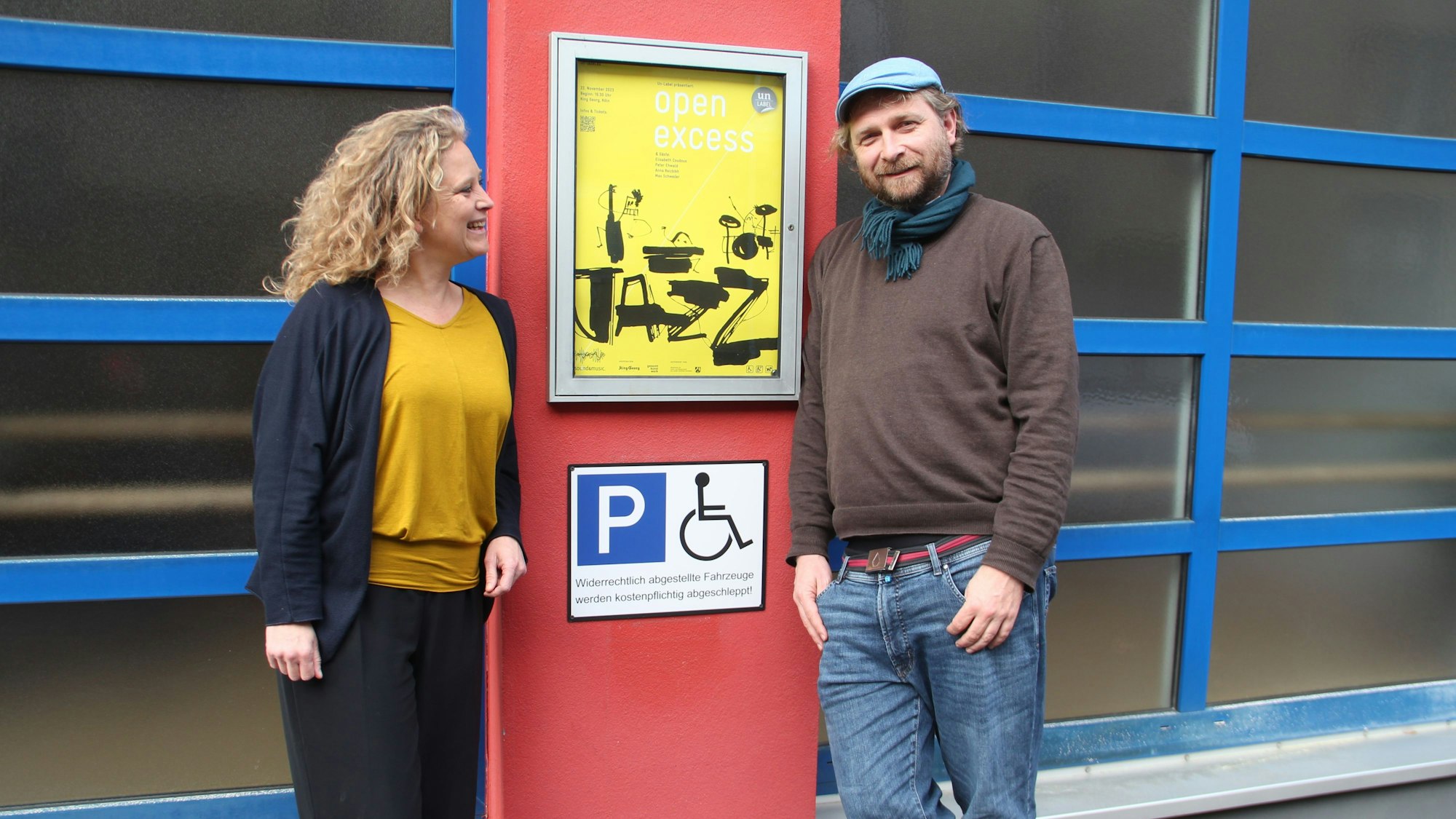 Eine Frau und ein Mann stehen neben einem Veranstaltungsschild und einem Piktogramm Behindertenparkplatz.