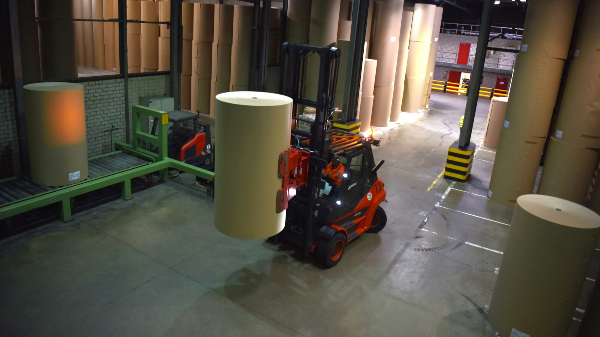 In einer Halle stehen große Papierrollen. Ein Gabelstapler transportiert eine Rolle von einem Rollband zu ihrem Platz in der Halle.