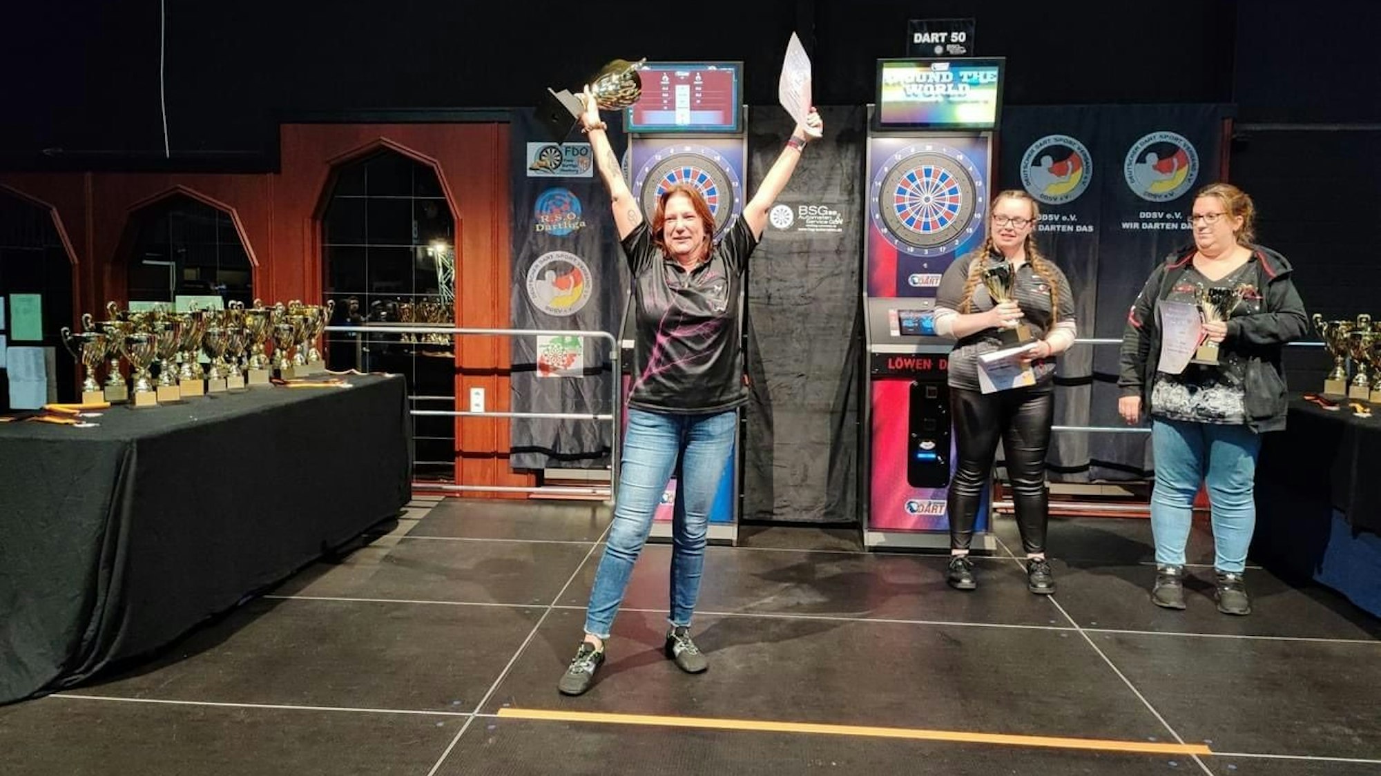 Eine Frau steht vor einem Dartbord und hält einen Pokal in den Händen. Die Arme hat sie zum Jubel ausgestreckt.