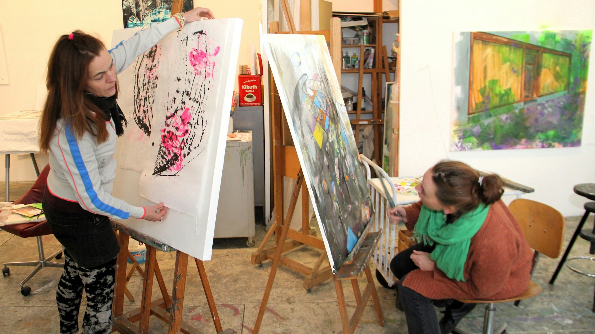 Zwei Frauen arbeiten in einem Atelier, eine steht vor einer Staffelei, die andere sitzt vor einer zweiten Staffelei.