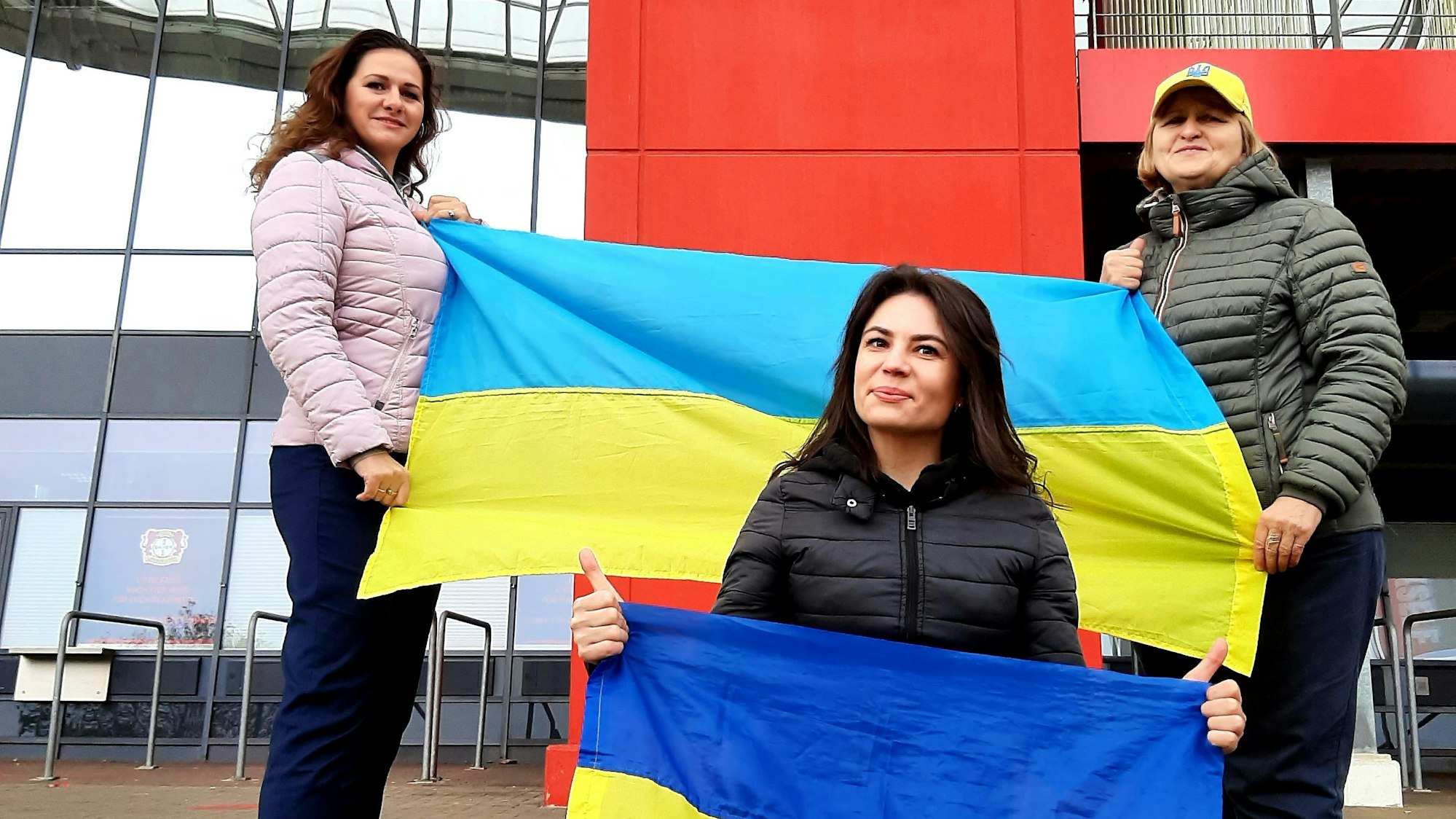 Die Ukrainerinnen (v.l.) Bilozin Liubov, Viktoriia Zhevlova und Nadiia Shepel stehen mit Ukraine-Flaggen vor der Bay-Arena, wo am kommenden Montag, 20. November,das Qualifikationsspiel zur Fußball-Europameisterschaft 2024 zwischen der Ukraine und Italien stattfindet.