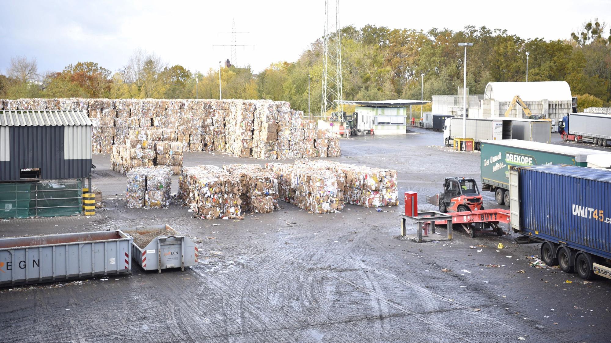 Auf einem Platz sind zusammengepresste Altpapierballen aufgestapelt, rechts im Bild sind Lkw zu erkennen.