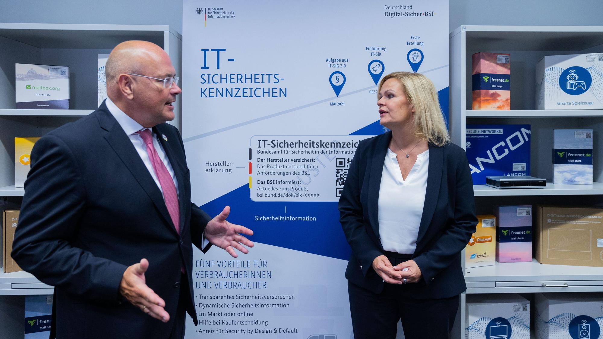 Arne Schönbohm gestikuliert mit beiden Händen in Richtung Nancy Faeser. Die beiden stehen vor Regalen und einem Plakat, auf dem „IT-Sicherheits-Kennzeichen“ steht.