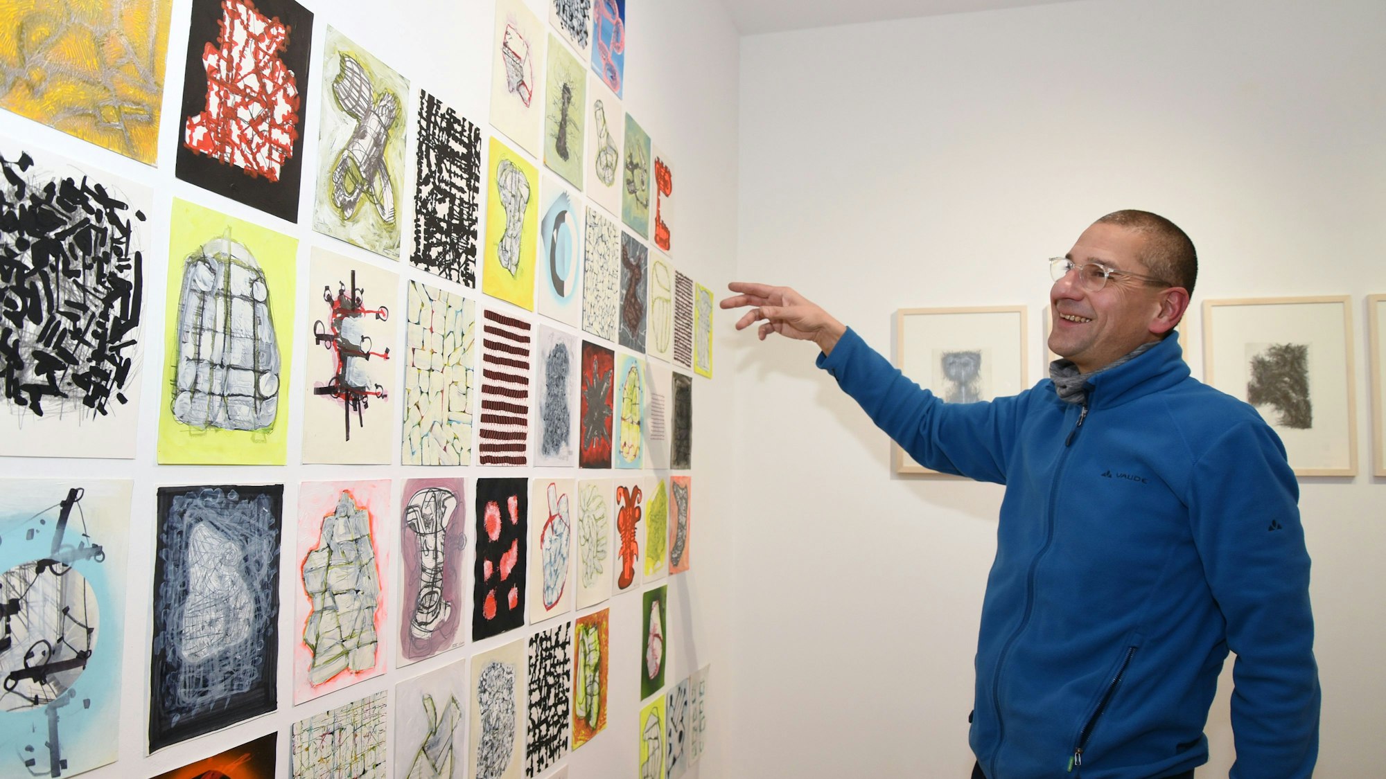 Ein Mann steht vor einer Wand mit vielen in Reihen angeordneten kleinformatigen Zeichnungen.