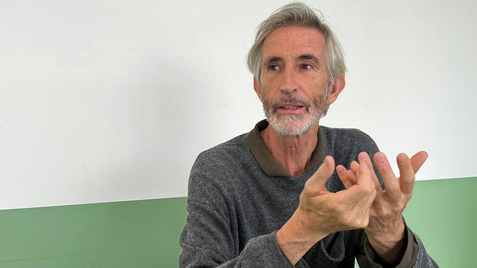 Der Künstler Francis Alÿs sitzt vor einer grün-weißen Wand.