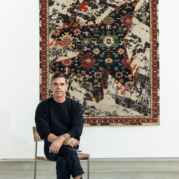 Jan Kath sitzt vor einem Teppichbild aus der Serie „Rug Bombs“.