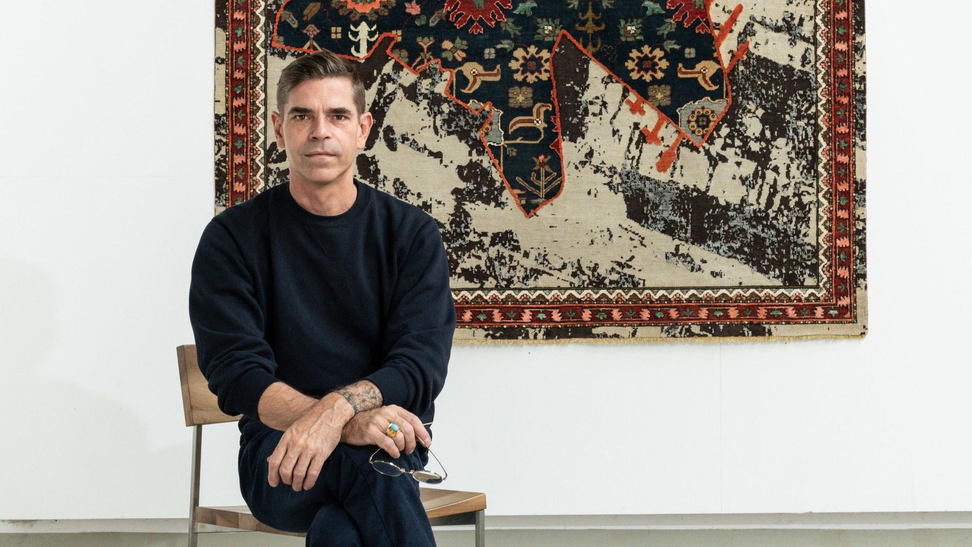 Jan Kath sitzt vor einem Teppichbild aus der Serie „Rug Bombs“.