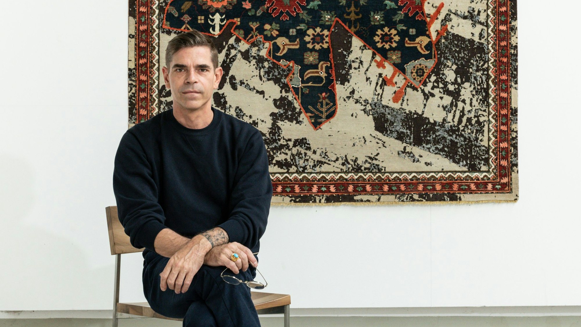 Jan Kath sitzt vor einem Teppichbild aus der Serie „Rug Bombs“.