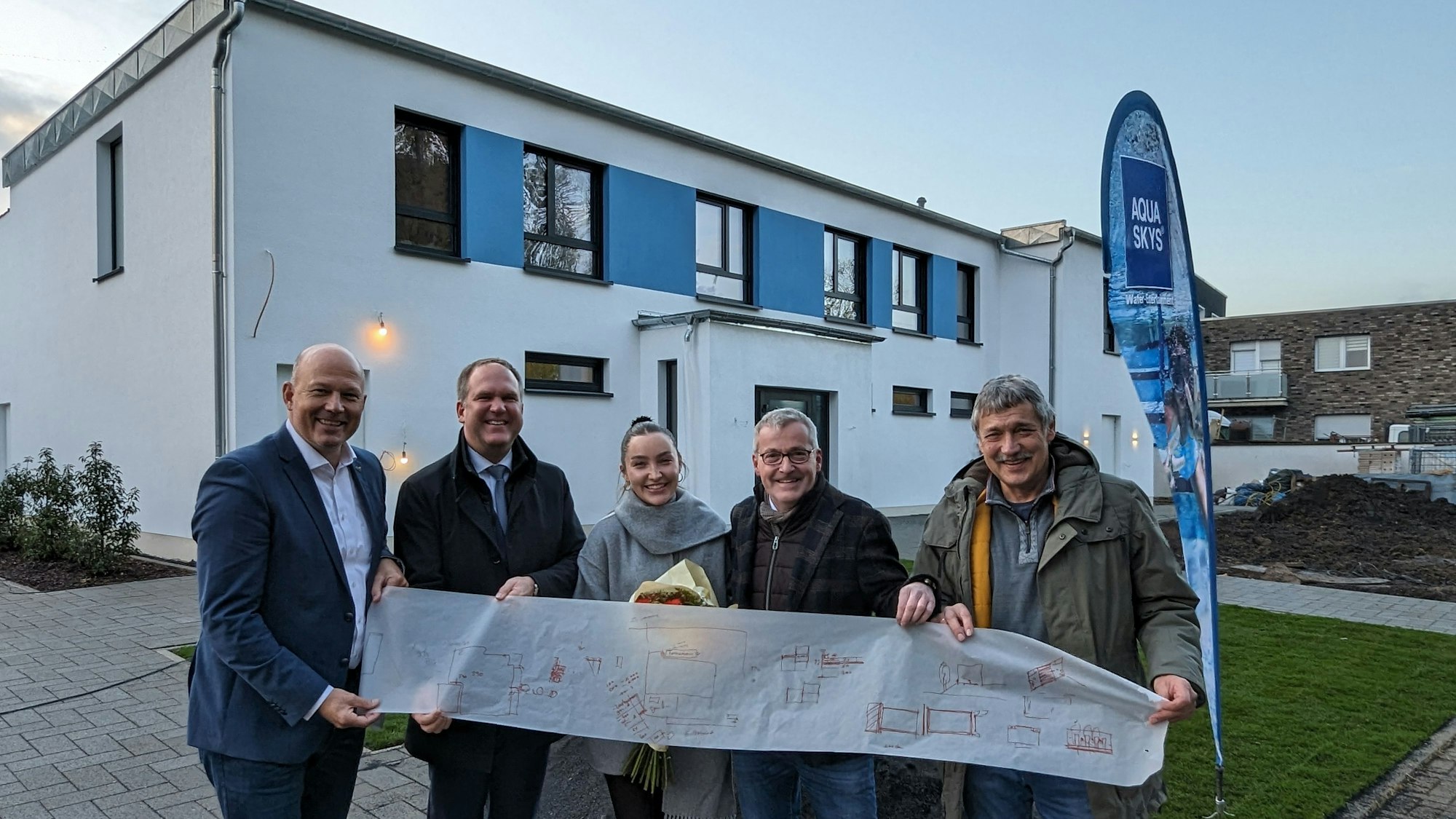 Eine Reihe von Menschen halten eine lange Pergamentpapierrolle vor einen zweigeschossigen Neubau.