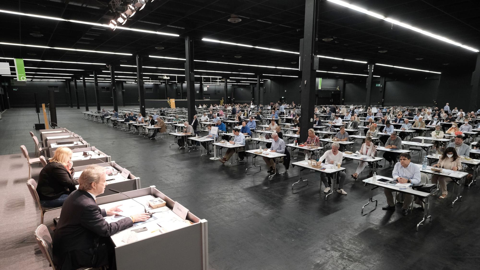 Blick in die Messehalle 11 bei einer Parteiveranstaltung. Vorne sitzen Menschen auf dem Podium. Im Hintergrund Teilnehmende an Einzeltischen. Die Decke und der Boden sind schwarz.