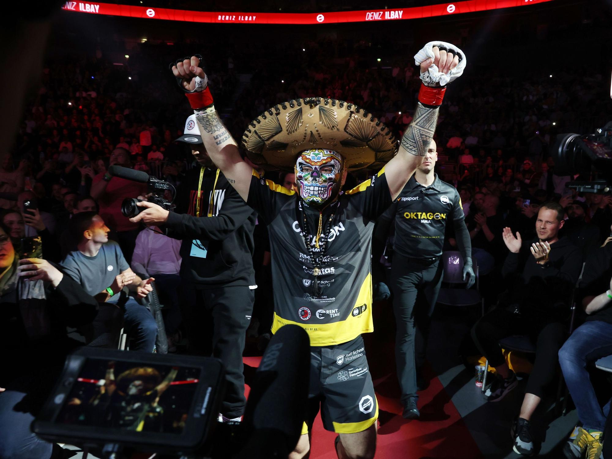 Deniz Ilbay mit Sombrero und mexikanischer Maske und erhobenen Armen.