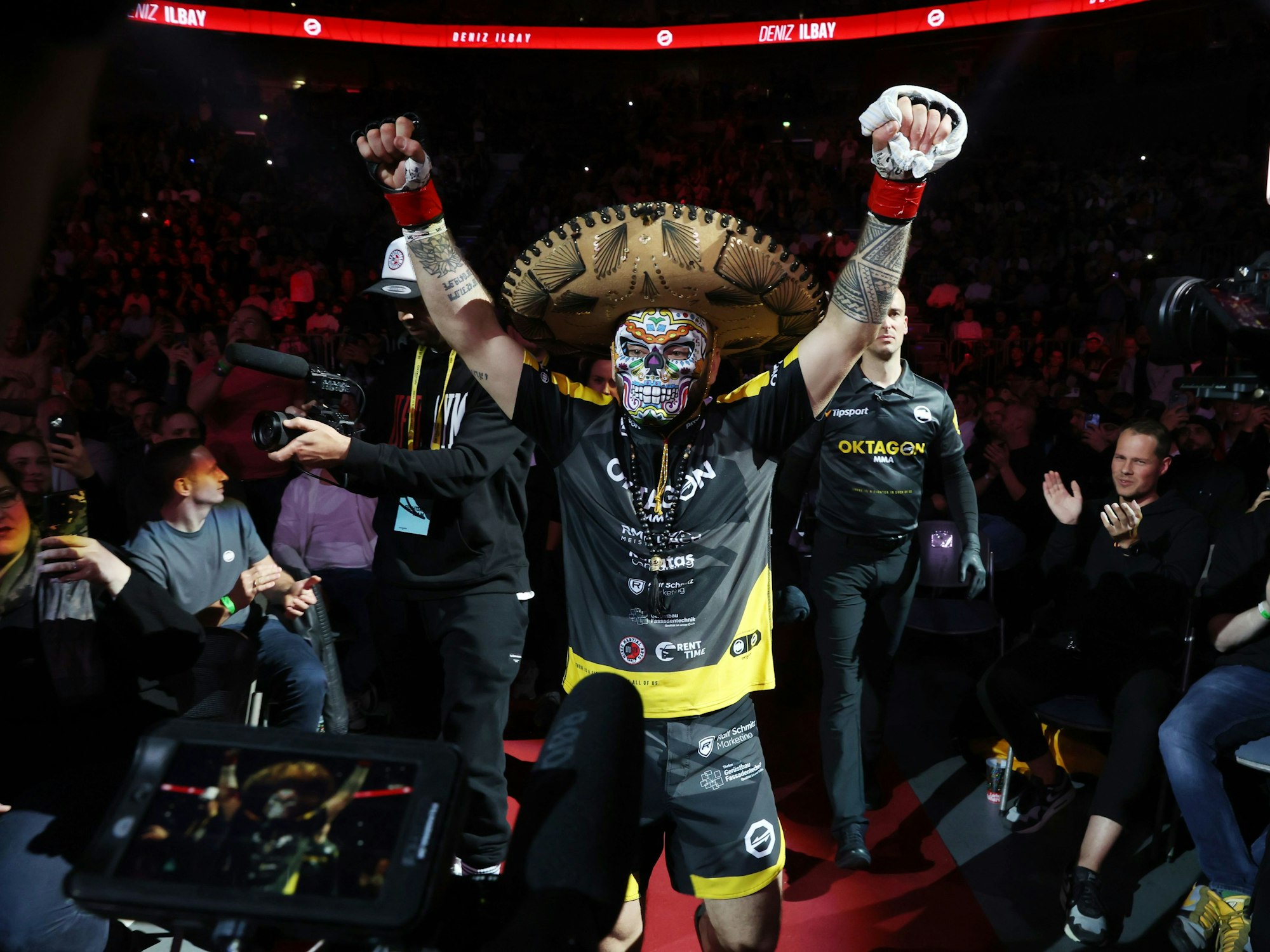 Deniz Ilbay mit Sombrero und mexikanischer Maske und erhobenen Armen.