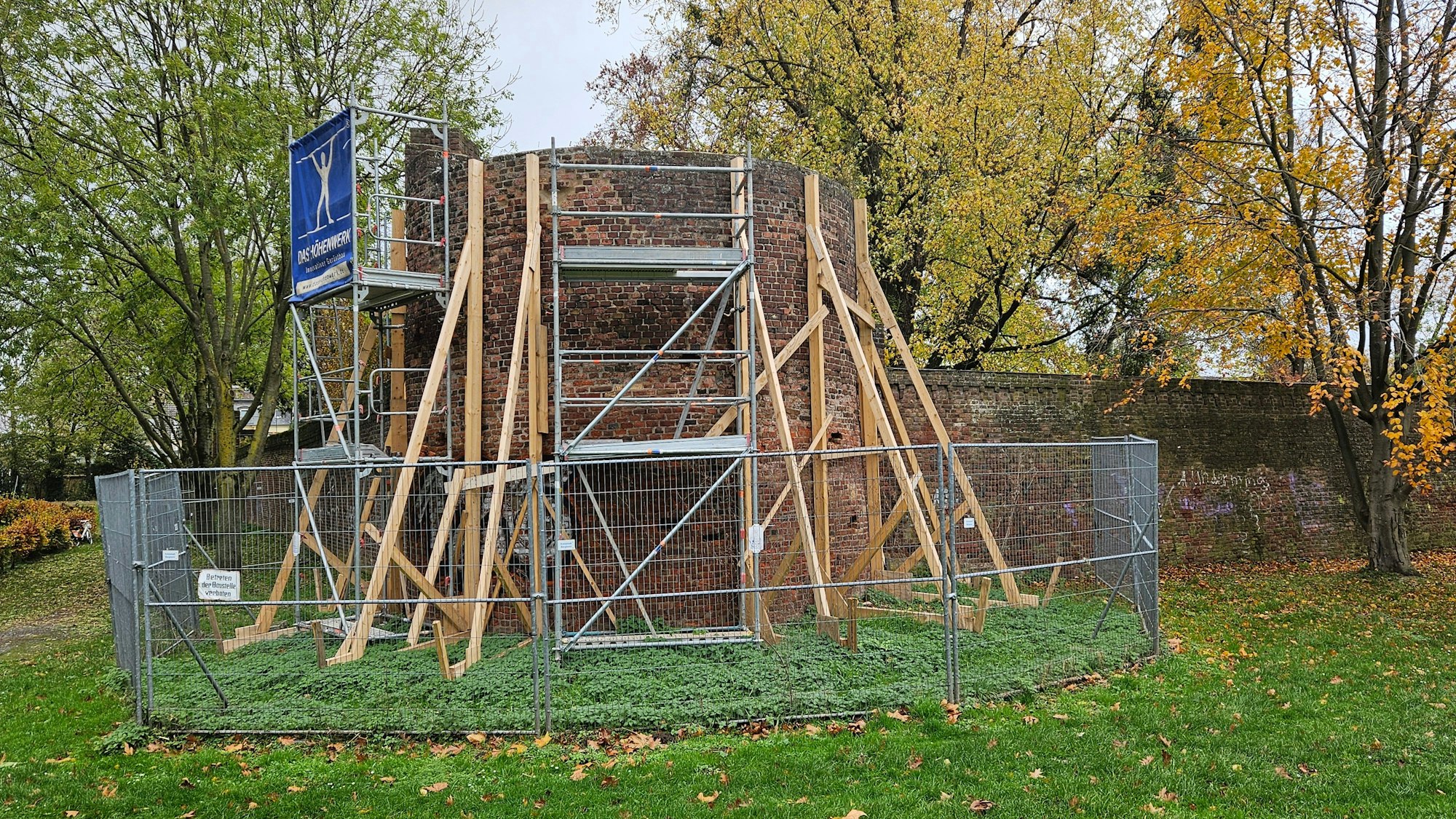 Kanthölzer, Gerüste und ein Bauzaun sichern einen Turm in einer Mauer.
