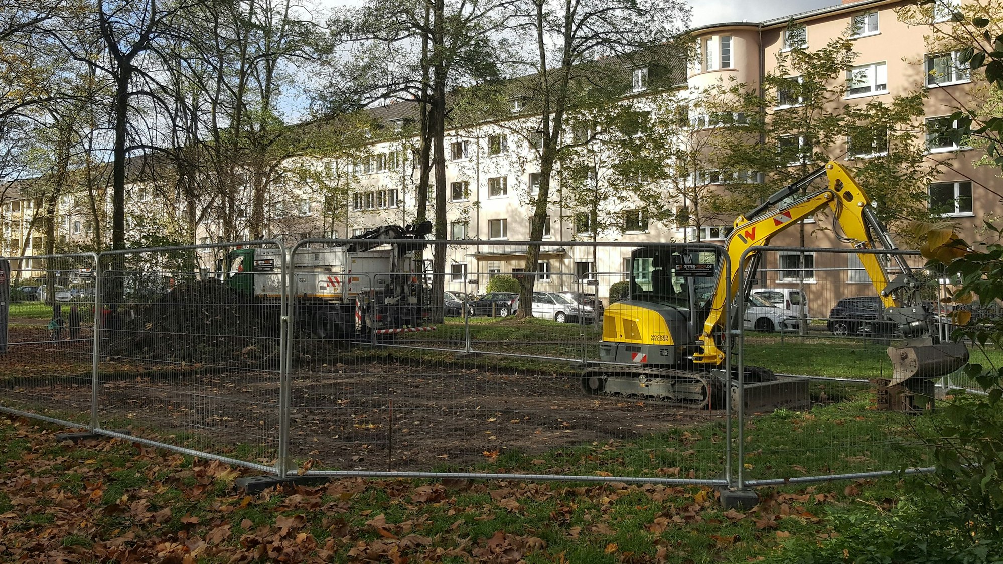 Ein Bagger steht in einer umzäunten Fläche inmitten einer größeren Grünfläche.