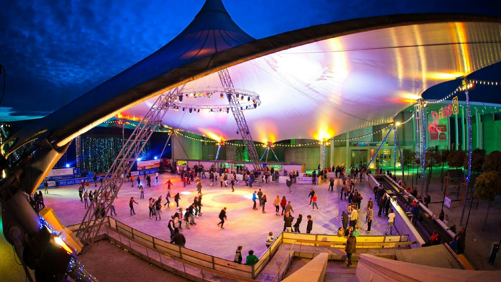 Menschen laufen Schlittschuh unter einem bunt beleuchteten Zeltdach.