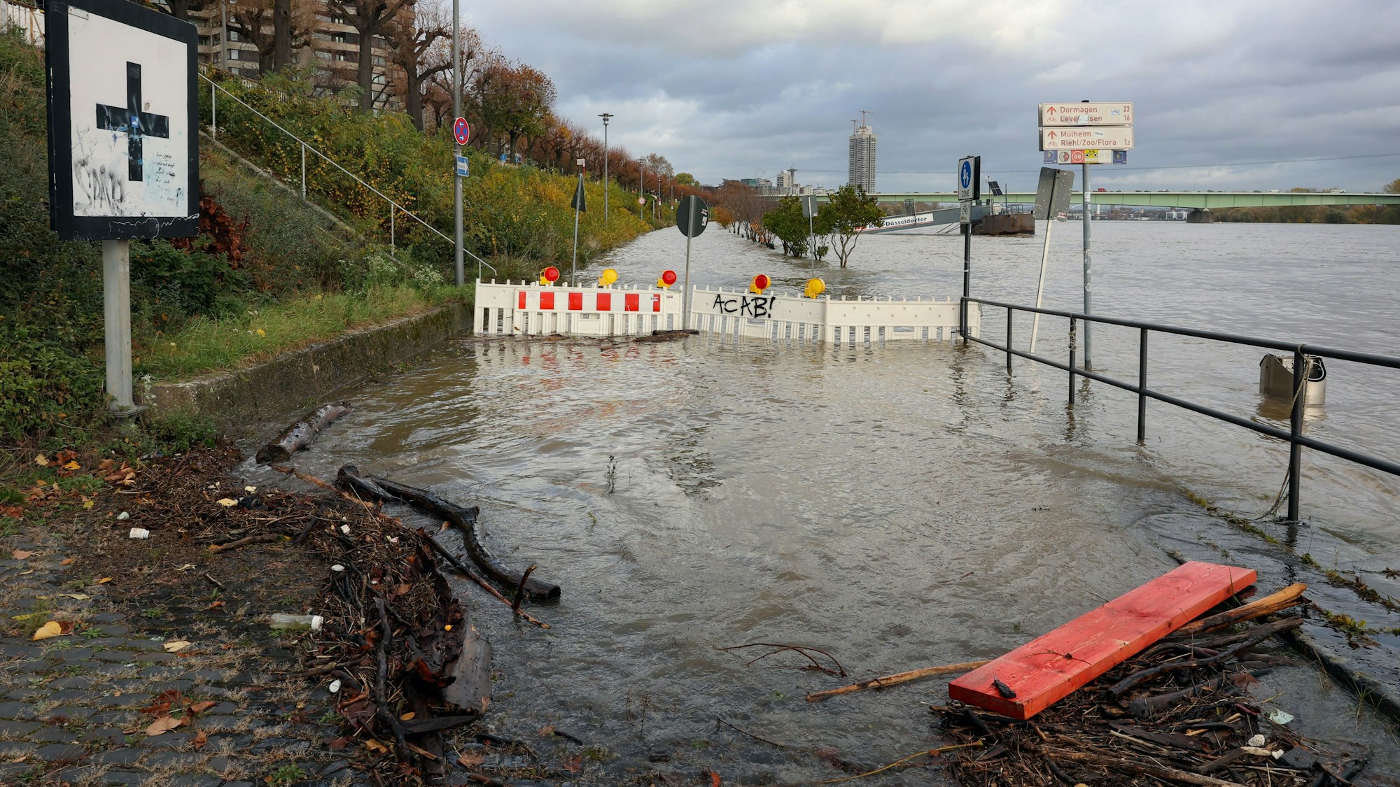 Blick auf das vom Wasser überspülte Rheinufer, im Wasser stehen Absperrbaken, rechts sind Geländer und Wegweiser zu sehen.