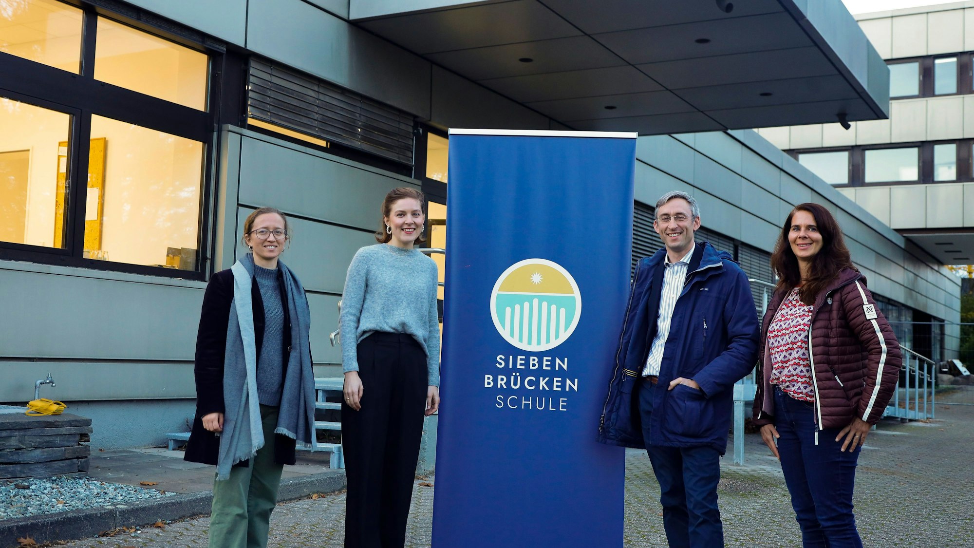 Vier Menschen stehen vor dem Logo einer Schule.