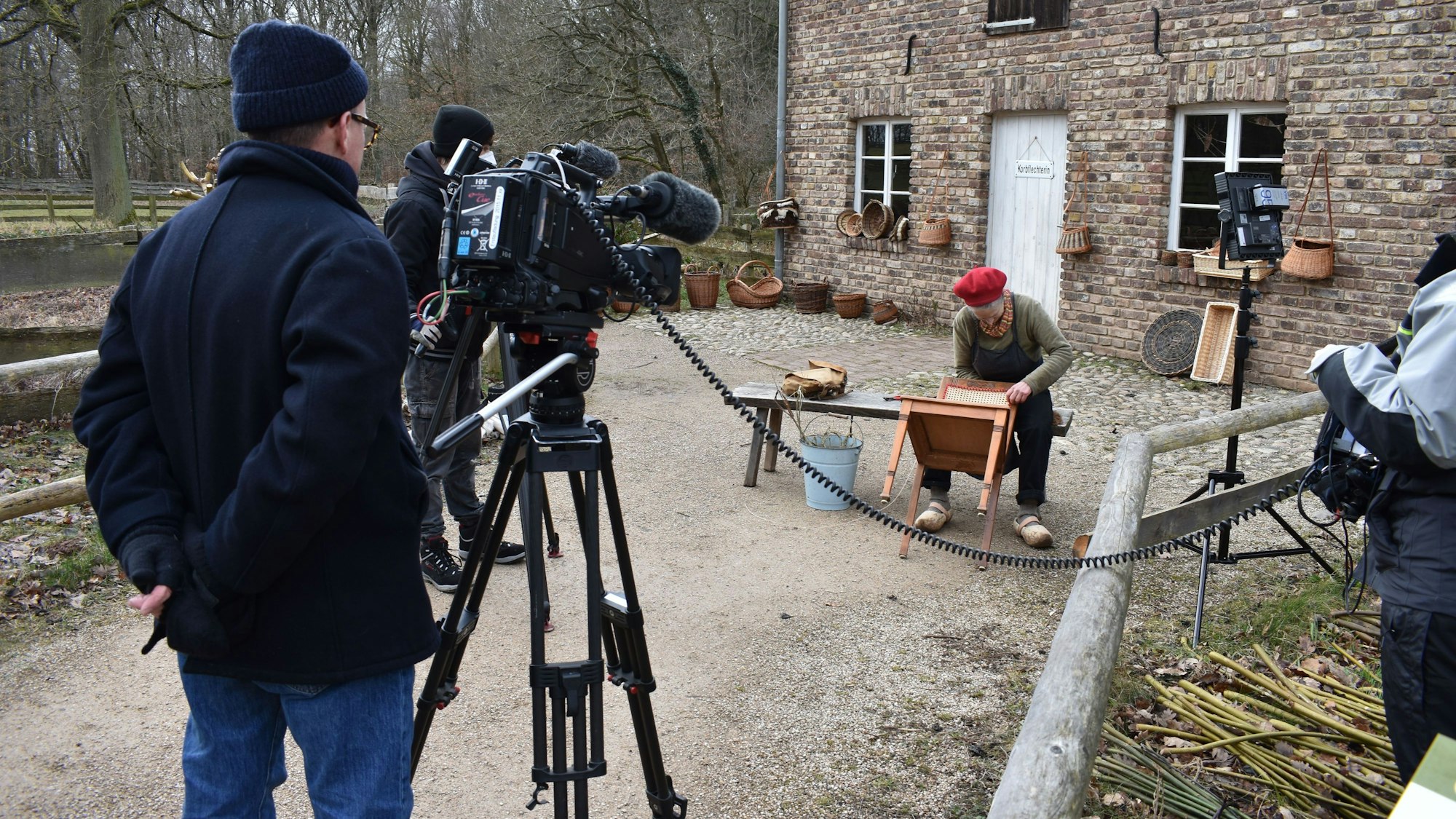 Ein Kamerateam filmt eine Korbflechterin, die im Freien vor der historischen Korbmacherwerkstatt in Kommern am Geflecht eines Stuhls arbeitet.
