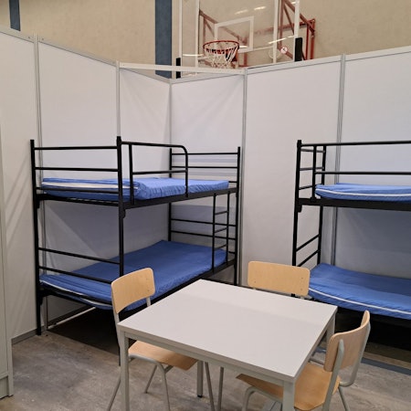 Zu sehen ist eine Wohnbox mit zwei Etagenbetten, einem Tisch mit Stühlen und einem Schrank. Die Wände sind aus Sperrholz, das mit Kunststoff (PVC) beschichtet ist.