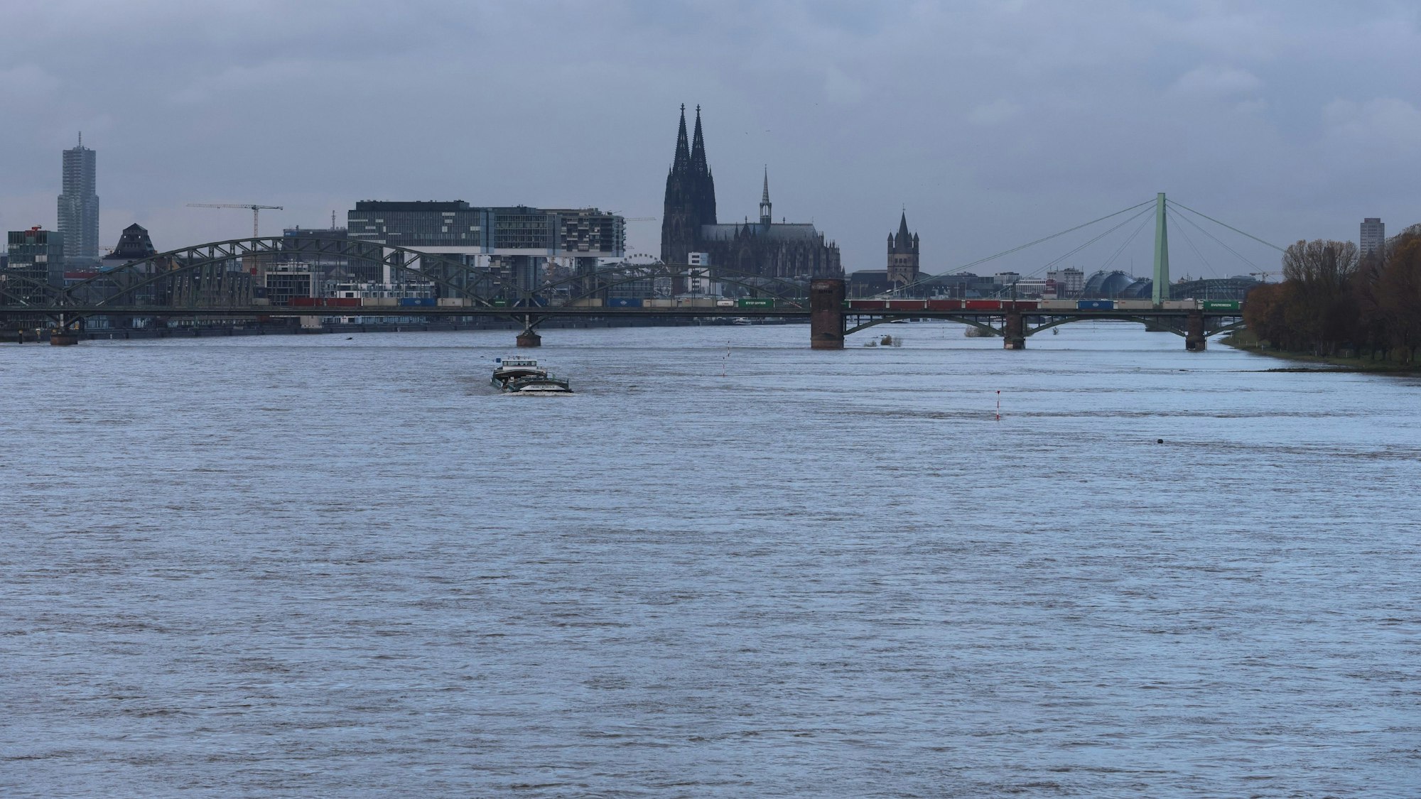Ein Frachtschiff ist auf dem Rhein unterwegs. Im Hintergrund sind die Südbrücke und das Stadtpanorama zu sehen. Die gesamte untere Bildhälfte ist vom Wasser eingenommen.