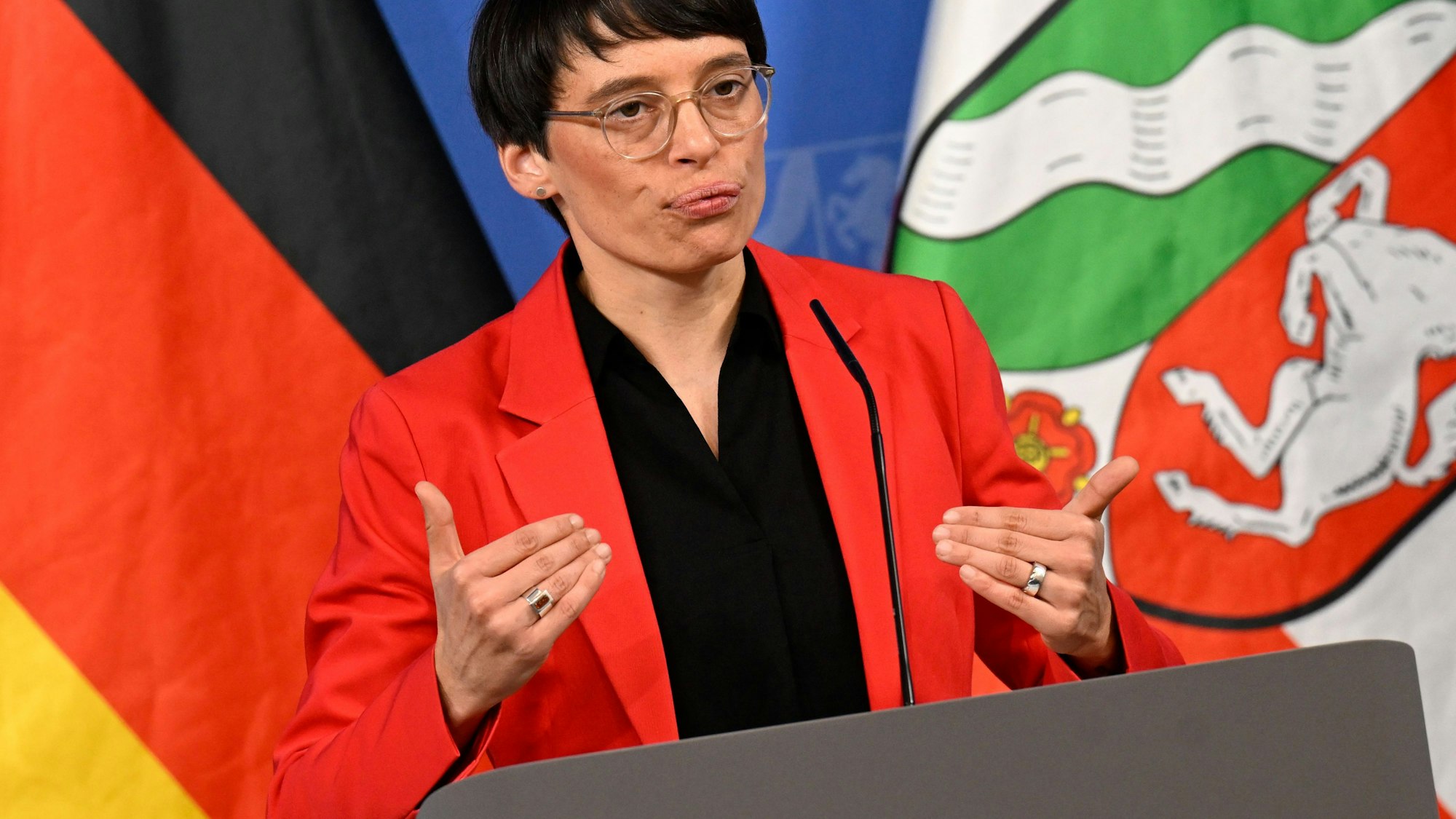 Josefine Paul, Integrationsministerin von Nordrhein-Westfalen, steht an einem Rednerpult. Hinter ihr zu sehen sind die deutsche und die nordrhein-westfälische Flagge.