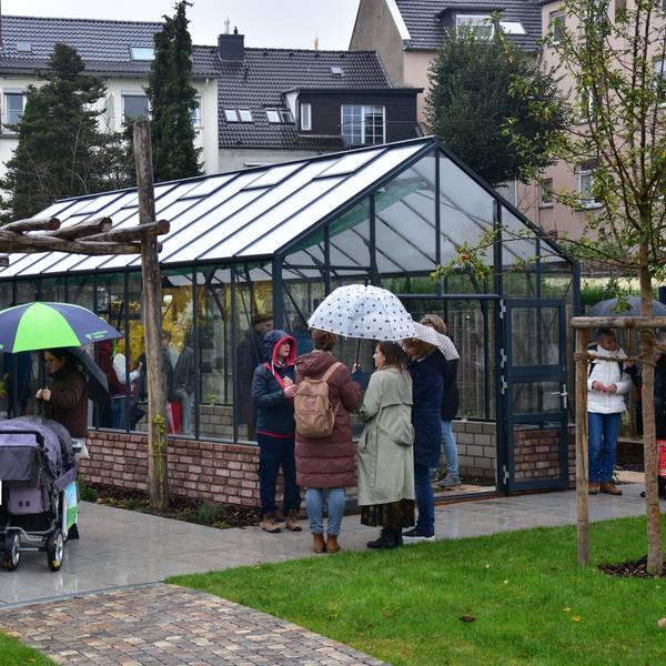 Leute mit Regenschirmen stehen vor Gewächshaus. Wohnhäuser im Hintergrund.