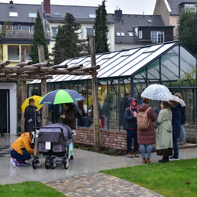 Leute mit Regenschirmen stehen vor Gewächshaus. Wohnhäuser im Hintergrund.