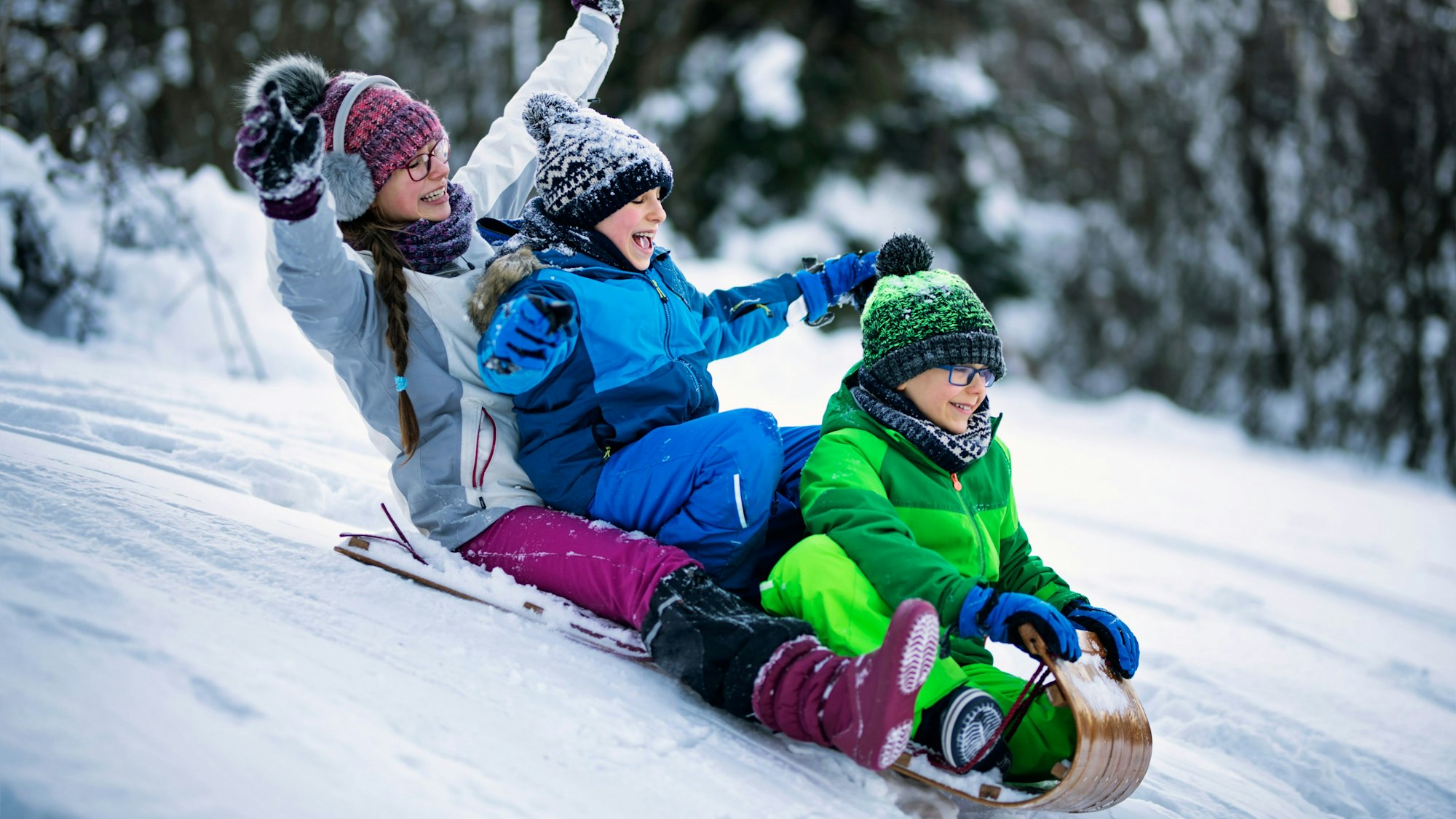 Drei Kinder rodeln auf einem Schlitten den Berg zusammen runter.