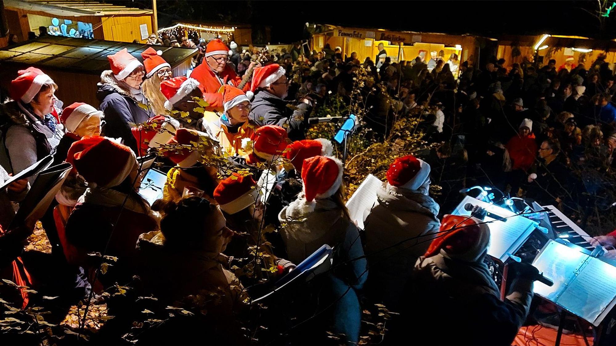 Menschen mit Nikolausmützen stehen im Dunkeln auf einem Weihnachtsmarkt und halten Noten in der Hand.
