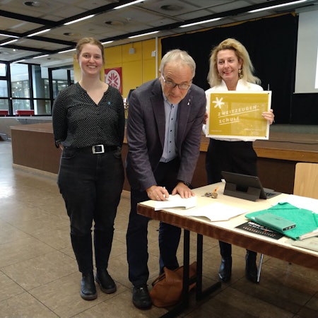 Zwei Frauen und ein Mann stehen an einem Tisch in einem Schulflur. Der Mann unterzeichnet gerade ein Schriftstück.