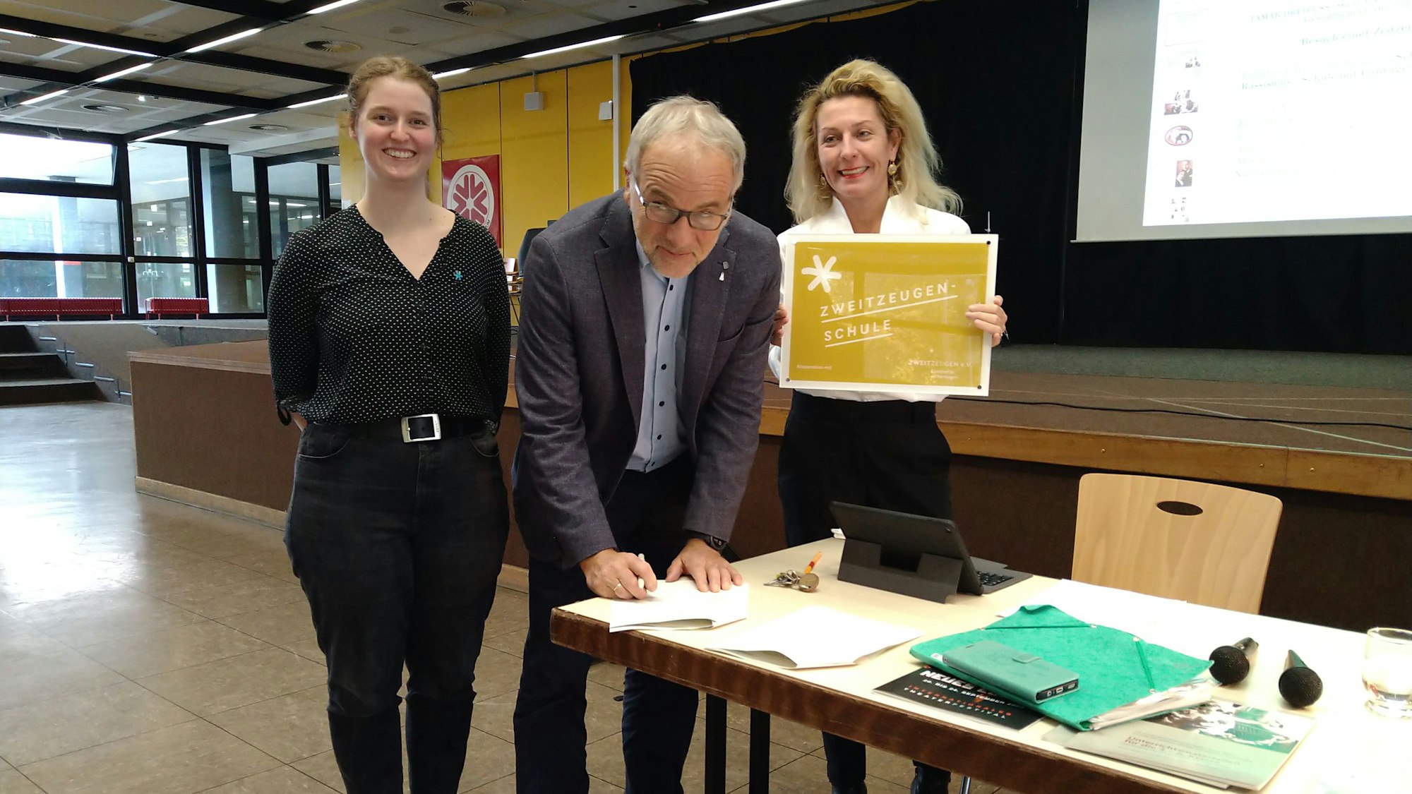 Zwei Frauen und ein Mann stehen an einem Tisch in einem Schulflur. Der Mann unterzeichnet gerade ein Schriftstück.