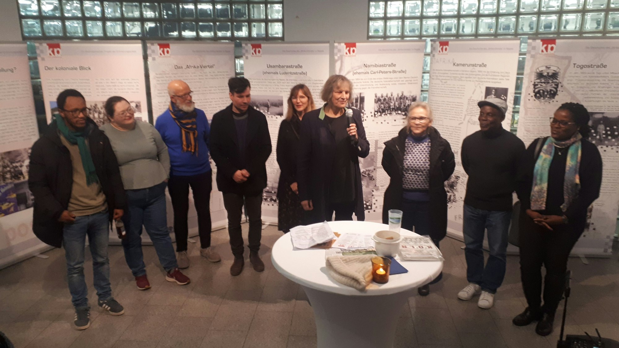 Vor Aufstellwänden stehen etwa zehn Menschen. Eine Dame hält ein Mikrofon in der Hand.