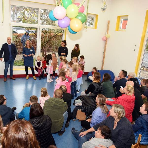 In einem großen Raum sind viele Kinder und Erwachsene unter bunten Luftballons versammelt.