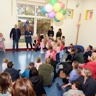 In einem großen Raum sind viele Kinder und Erwachsene unter bunten Luftballons versammelt.