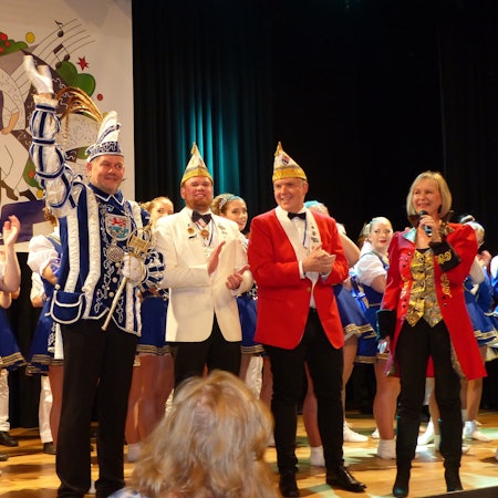 Auf einer Bühne stehen Mitglieder einer Karnevalsgesellschaft. In ihrer Mitte steht der Prinz und winkt ins Publikum.