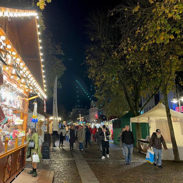Weihnachtsmarktbuden aus Holz mit geschmückten Dächern reichen sich über den Marktplatz. Menschen schlendern zwischen ihnen umher.
