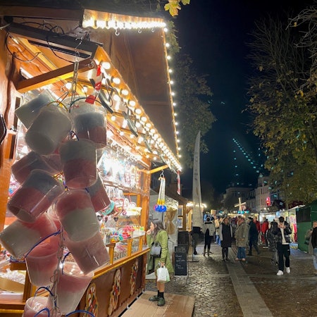 Weihnachtsmarktbuden aus Holz mit geschmückten Dächern reichen sich über den Marktplatz. Menschen schlendern zwischen ihnen umher.